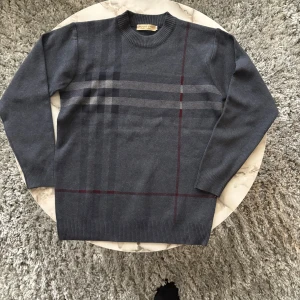 Burberry stickad tröja - Snygg grå stickad tröja från Burberry med klassiskt rutmönster i mörkgrått och vinrött. Rund halsringning och långa ärmar med ribbade muddar. Perfekt för dig som gillar stilrena och tidlösa plagg med en touch av lyx. Otroligt skön att ha på sig och väldigt bra passform, helt ärligt 10/10 sweatshirt