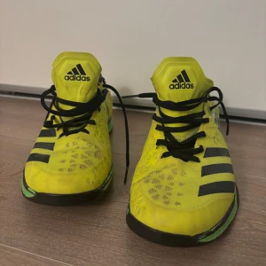 Adidas innebandy/inomhusskor - Säljer ett par Adidas sneakers i en riktigt cool gul färg med svarta ränder och detaljer. Skorna har grön sula och svart insida, samt snörning framtill. Ovandelen är i mesh för bra ventilation och lätt känsla. Perfekta för dig som gillar sportig stil och sticker ut.