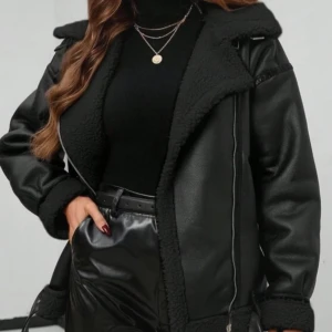 Svart teddyjacka med skinnlook - Snygg svart teddyjacka med skinnlook och bred krage. Jackan har detaljer i fuskpäls, dragkedja och bältesdetaljer vid nederkant och ärmslut. Oversized passform och långärmad, perfekt för dig som gillar en cool och trendig stil.