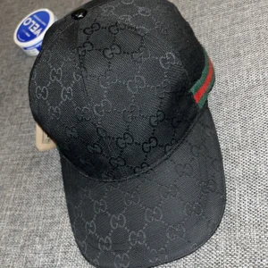 Gucci keps med GG-mönster - ”ÄKTAA”.   Säljer en svart keps från Gucci med klassiskt GG-monogram över hela kepsen. Den har en böjd skärm och justerbar rem baktill. På sidan finns en ikonisk grön och röd rand. Materialet är slitstark bomull och kepsen är tillverkad i Italien.