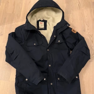 Marinblå vinterjacka  från Fjällräven - Vinterjacka från Fjällräven i marinblått med mysigt vitt fleecefoder och huva. Jackan har flera fickor med knappar, dragkedja och tryckknappar framtill samt klassisk Fjällräven-logga på ärmen. Perfekt för kalla dagar och outdoor vibes. Modellen heter Greenland och nypris ligger runt 3500kr. Hör av er vid frågor!