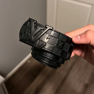 Svart Louis Vuitton rutigt bälte - Säljer ett svart bälte från Louis Vuitton med klassiskt rutigt mönster och stor LV-logga i metall framtill. Bältet är gjort i skinn och har en bred, stilren design som sticker ut. Perfekt accessoar för att lyfta din outfit.