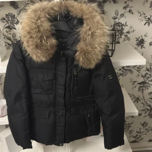 Svart pufferjacka med päls från Hollies - Svart pufferjacka från Hollies med stor fluffig päls runt huvan. Jackan har flera fickor med dragkedja och knappstängning, samt snygga detaljer på ärmarna. Perfekt för kalla dagar med sin tjocka och värmande design. på ena fickan så saknas bandet till dragkedjan, Det är de svarta bandet som inte finns med och dragkedjan i mitten. men funkar hur bra som helst.