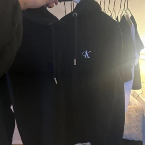 Svart hoodie från Calvin Klein Jeans - Svart hoodie från Calvin Klein Jeans med broderad CK-logga på bröstet. Tröjan har huva med snörning och långa ärmar.. Klassisk och stilren design som funkar till allt.
