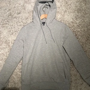 Grå hoodie från Barbour med broderi - Snygg grå hoodie från Barbour med broderad logga på bröstet. Tröjan har huva med snörning, ribbade muddar och en klassisk loose passform. Perfekt för chill dagar och enkel att matcha med jeans eller joggers.