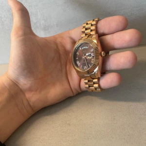 Guldfärgad klocka från Nibosi - Snygg guldfärgad klocka från Nibosi med metallarmband och mörkbrun urtavla. Klockan har romerska siffror, dag- och datumvisning samt en klassisk rund form. Länken är robust och ger en lyxig känsla. Perfekt för dig som vill sticka ut med stil. Men ända är att batteriet har stannat så de behövs bytas annars bra skick skicka samma dag eller dagen efter😊