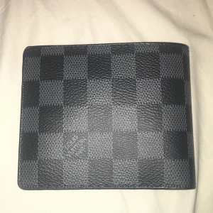  rutig svart plånbok - Snygg plånbok  i klassiskt rutigt mönster Damier Graphite. Svart färg med flera kortfack och fönster för ID. Tillverkad i slitstarkt skinn med lyxig känsla och stilren design. Perfekt för dig som vill ha något exklusivt i fickan.
