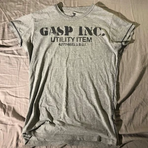 Grå GASP INC. t-shirt utility style - Snygg grå t-shirt från GASP INC. med utility-tryck på bröstet och mindre logga på ryggen. T-shirten har korta ärmar, rund hals och kontrastsömmar vid ärmslut och hals. Perfekt för dig som gillar sportig och urban stil.