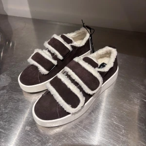 Snygga Snekers - Säljer ett par mörkbruna sneakers från Duffy i storlek 39. Skorna har vit fluffig fuskpäls på insidan och runt kardborrebanden, samt en vit platt sula. Utsidan är i mocka. Perfekta nu till vintern med ett par snygga och varma sneakers💕Helt nya, pga att de var för stora för mig. Skickas i den kartongen jag fick de i. 
