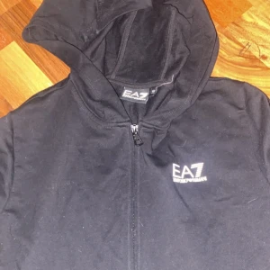Svart hoodiejacka EA7 Emporio Armani - Svart hoodiejacka från EA7 Emporio Armani med dragkedja framtill och logga på bröstet. Jackan har huva och är tillverkad i mjukt bomullsmaterial, perfekt för en chill och sportig look. Passar dig som gillar stilrena och enkla plagg med premiumkänsla.