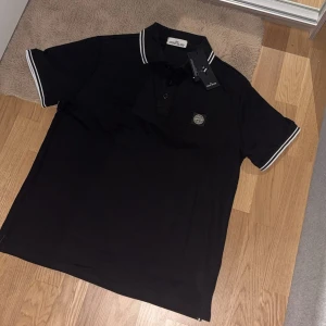 Svart pikétröja från Stone Island - Svart pikétröja från Stone Island med klassisk krage och vita ränder på ärmslut och krage. Tröjan har knappslå framtill och ikonisk Stone Island-logga på bröstet. Tillverkad i bomull för en skön och stilren känsla.