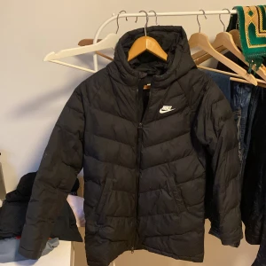 Svart pufferjacka från Nike - Svart pufferjacka från Nike med huva och vit logga på bröstet. Jackan har dragkedja framtill och två sidofickor. Den är quiltad och har en sportig look som passar perfekt när det är kallt ute.