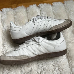 Adidas Samba vita sneakers i skinn - Klassiska Adidas Samba sneakers i vitt skinn med grå mockadetaljer och bruna gummisulor. Skorna har de ikoniska tre ränderna på sidorna, snörning och blå Adidas-logga på plösen. Perfekta för dig som gillar retrostil och streetwear.