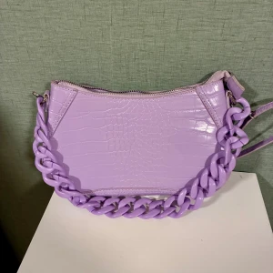 Lila axelväska från Bershka - Superfin lila axelväska från Bershka med croco-mönstrad yta och chunky plastkedja som handtag. Väskan har dragkedja upptill och är gjord i syntetmaterial. Perfekta färgklicken för att pigga upp en lite tråkigare outfit 🙌