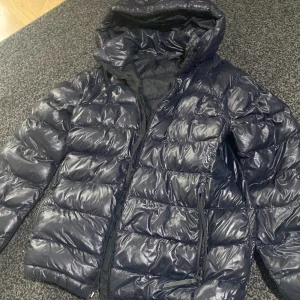 Mörkblå Peak Performance dunjacka - Säljer en mörkblå dunjacka från Peak Performance med glansig yta och huva. Jackan har dragkedja framtill och på fickorna, samt ett snyggt logomärke på ärmen. Perfekt för kalla dagar och har en riktigt schysst puffer-look. Dock har dragkedjan lossnat men det går att byta ut. Köptes för 2000kr fast eftersom dragkedjan lossnat så säljs den för 400