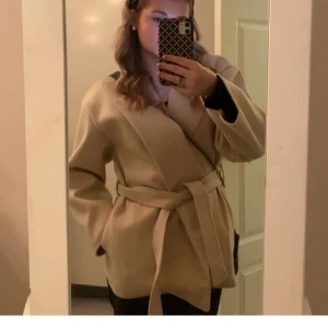 Beige omlott kappa med bälte - Säljer en beige kappa med omlott-design och bälte i midjan. Jackan har en stilren look med breda slag och långärmade ärmar. Perfekt för dig som gillar minimalistisk och clean stil. Materialet känns mjukt och har en snygg fall.