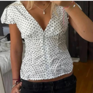 Vit prickig blus med v-ringning - Säljer en vit blus med svarta prickar och djup v-ringning. Blusen har korta puffärmar, knappar framtill och rynkade detaljer längs sidorna som ger en snygg siluett. Super fint med polka dots!!