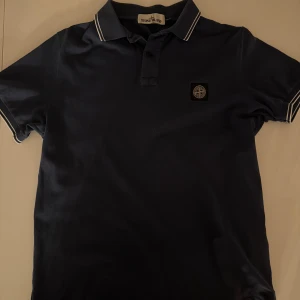 Marinblå pikétröja Stone Island - Snygg marinblå pikétröja från Stone Island med klassisk krage och vita ränder på krage och ärmslut. Tröjan har korta ärmar och ikonisk patch på bröstet. Perfekt för dig som gillar stilrena och sportiga plagg. Använd 2 gånger då storleken inte passade. Nypris 2100 pris kan diskuteras vid snabb affär!! 