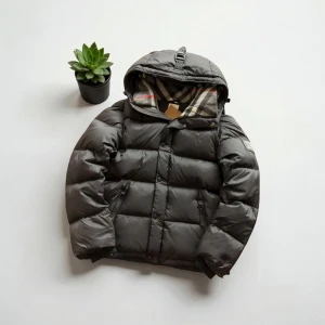 Svart pufferjacka från Burberry - Svart pufferjacka från Burberry med klassiskt rutigt foder i huvan och snygga detaljer som snörning och dragkedja. Jackan har en stor huva med spänne, två fickor med dragkedja och quiltad design. Perfekt för kalla dagar med sin fluffiga och stilrena look.