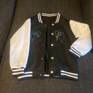 Svart och vit bomberjacka med tryck - Snygg svart och vit bomberjacka med coolt spelkontroll-tryck på bröstet. Jackan har vita ärmar, svarta muddar med vita ränder och knappar framtill. Perfekt för dig som gillar gaming och streetstyle.