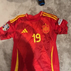 Spanien fotbollströja  - Röd fotbollströja från Adidas med Spaniens landslagsemblem, nummer 19 och Lamine Yamal på ryggen. Tröjan har gula detaljer, korta ärmar och officiella turneringsmärken på båda ärmarna. Tillverkad i lätt och ventilerande material, perfekt för fotbollsfans.