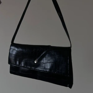 Svart axelväska i krokopräglat skinn - Stilren svart axelväska med krokopräglat mönster och silverfärgad knappstängning. Väskan har en avlång form och justerbar axelrem. Tillverkad i syntetmaterial med skinnlook, perfekt för dig som gillar trendiga accessoarer.