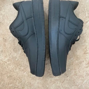 Nike Air Force 1 helsvarta sneakers - Säljer ett par helsvarta Nike Air Force 1 sneakers i klassisk low-modell. Skorna har en stilren design med rund tå, snörning och platt sula. Tillverkade i skinn med diskret Nike-logga på sidan och AIR-detalj på sulan. Perfekta för dig som gillar en clean och tidlös look.