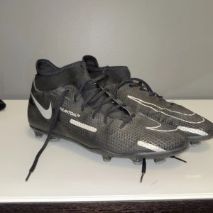 Nike Phantom GT2 Svarta Fotbollsskor - Svarta Nike Phantom GT2 fotbollsskor med vita detaljer och snörning. Skorna har en stickad ovandel i syntetmaterial och en högre strumpliknande krage för extra stöd. Perfekta för dig som vill ha grym kontroll på planen.