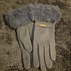Grå handskar med pälsdetalj - Snygga grå handskar med fluffig pälsdetalj vid handleden och en liten metallbricka med texten 'FASHION'. Handskarna har en stilren design och är tillverkade i mjukt syntetmaterial med pälsimitation upptill. Perfekta för kalla dagar när du vill vara både varm och trendig.