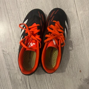 Adidas Predator fotbollsskor svart/röd - Säljer ett par Adidas Predator fotbollsskor i svart med röda och vita detaljer. Skorna har snörning, röda skosnören och innersula, samt mönstrad yttersula med dobbar för bra grepp på planen. Perfekta för dig som vill sticka ut på fotbollen!
