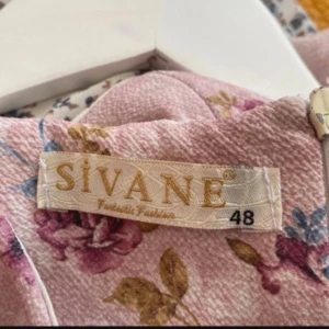 Blommig långklänning från Sivane - Långklänning i ljusrosa nyans från Sivane med vackert blommigt mönster i lila, blått och grönt. Klänningen har lång ärm, rund halsringning och plisserad nederdel som ger en romantisk känsla. Perfekt för dig som gillar feminina och mjuka toner.