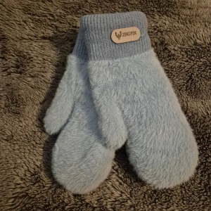 Ljusblå fluffiga vantar - Supermysiga ljusblå vantar från Jingpin med fluffig utsida och mjukt, beige foder. Vantarna har en ribbstickad mudd och en snygg logodetalj i trä. Perfekta för kalla vinterdagar när du vill hålla händerna varma och stiliga.