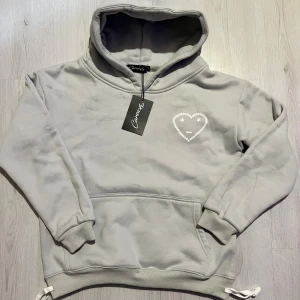 Grå hoodie med hjärtdetalj Carrsicko - Snygg grå hoodie från Carrsicko i mjuk bomull med huva och ribbade muddar. På bröstet finns ett vitt hjärta med ansikte och stjärnor som ögon. Tröjan har snörning i nederkant och en klassisk känguruficka framtill.