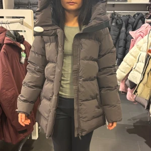 Mörkgrå pufferjacka med huva - Säljer en mörkgrå pufferjacka med stor huva och dragkedja framtill. Jackan har en quiltad design med breda kanaler och är långärmad. Perfekt för kalla dagar när du vill hålla dig varm och samtidigt se chill ut.