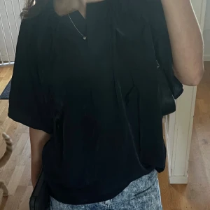 Svart blus med korta ärmar - Säljer en svart blus med korta ärmar och avslappnad passform. Blusen har en enkel design med lätt glansigt material och en liten v-ringning vid halsen. Perfekt att matcha med jeans eller kjol för en stilren look. Använt fåtal gånger och den är i nyskick 
