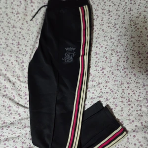 Svarta Messi mjukisbyxor med ränder - Ett par svarta träningsbyxor med dragsko i midjan. Byxorna har tre ränder i rosa och beige längs sidan och en broderad logotyp på vänster ben.