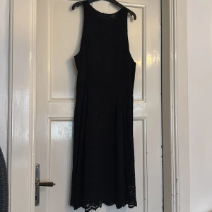 Svart spetsklänning från Lindex - Svart ärmlös klänning från Lindex med vacker spets och rund hals. Klänningen har en lätt utställd form och knälång längd med dekorativ kant nedtill. Dragkedja i ryggen för enkel på- och avtagning. Perfekt för dig som gillar stilren och elegant look.