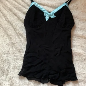 Svarta shorts med blå rosett från Divided - Säljer ett par svarta shorts med inbyggd topp från Divided. De har tunna axelband och en ljusblå satinrosett framtill, samt matchande blå kant vid urringningen. Materialet är lätt och flowy, perfekt för chill dagar. Supersöt detalj med rosetten!