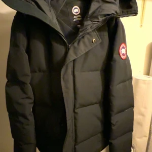 Macmillan Parka Black - Stilren och varm Canada Goose Macmillan Parka i klassisk svart färg. Jackan har dunfyllning av högsta kvalitet som ger utmärkt värme och komfort även under de kallaste vinterdagarna. Modern, något kortare passform med justerbar huva, dolda dragkedjor och praktiska fickor. Den ikoniska Canada Goose-logotypen sitter på ärmen och ger jackan ett exklusivt uttrycker 