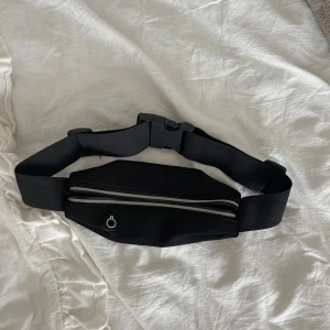 Svart midjeväska med dragkedja - Snygg svart midjeväska med justerbart band och smidig plastspänne. Väskan har en silverfärgad dragkedja och ett litet hål för hörlurar. Perfekt för att ha mobilen och nycklar nära till hands när du är på språng.