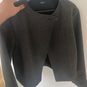 Mörkgrå kavajjacka från Gina Tricot - Snygg mörkgrå kavajjacka från Gina Tricot i stilren design med rund halsringning och asymmetrisk knäppning med knapp. Jackan har lång ärm och är tillverkad i ett ulligt material som ger en exklusiv känsla. Perfekt för dig som gillar minimalistisk stil. Aldrig använd men prislapp finns ej kvar.