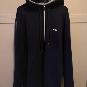 Mörkblå hoodie med dragkedja från BOSS - Snygg mörkblå hoodie från BOSS med vit dragkedja och huva. Tröjan har långa ärmar, snörning i huvan och diskret BOSS-logga på bröstet. Perfekt för en chill och stilren look.