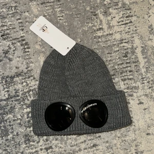 Grå C.P. Company mössa med goggles - Säljer en grå mössa från C.P. Company med ikoniska svarta goggles på uppviket. Mössan är ribbstickad och har en snygg, urban look som sticker ut. Har kvitto också 