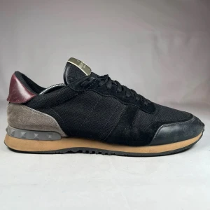 Valentino Garavani Rockrunner sneakers - Svarta Valentino Garavani Rockrunner sneakers med mesh och mocka på ovandelen, grå och vinröda detaljer vid hälen samt beige mellansula. Snygga nitar på bakre delen av sulan och klassisk snörning. Perfekt för dig som gillar exklusiva och stilrena sneakers.