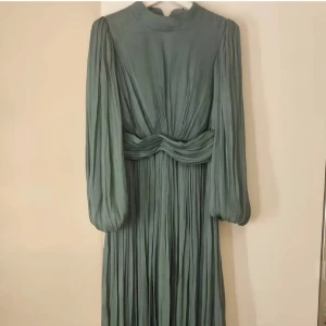 Lång grön plisserad klänning med markerad midja - Säljer en lång, plisserad klänning i dämpad grön nyans med höga puffärmar och markerad midja. Klänningen har hög halsringning, vackra draperingar och knytning i ryggen. Materialet är lätt och ger ett elegant fall. Perfekt för dig som gillar en stilren och sofistikerad look.