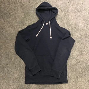 Mörkblå hoodie från Pier One - Snygg mörkblå hoodie från Pier One med klassisk huva och vita snören. Tröjan har en stor magficka och ribbade muddar vid ärmslut och nederkant. Perfekt för chill dagar och enkel att matcha med jeans eller joggers.