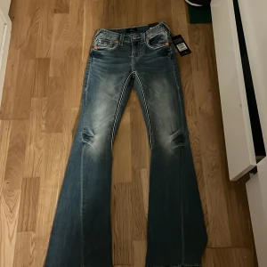 True Religion Joey Ultra Flare Jeans - Snygga blå jeans från True Religion, modell Joey Ultra Flare med coola slitningar och kontrastsömmar. Midwaist passform och riktigt breda ben för en statement look. Klassiska fickor med knappar och orange detaljer framtill. Perfekt för dig som gillar retrovibbar och unik stil. Pris kan diskuteras