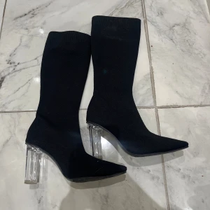 Svarta stövletter med transparent klack - Säljer ett par svarta stövletter med stickad textur och spetsig tå. Skorna har en hög, transparent klack som ger en modern och edgy vibe. Perfekta för dig som vill sticka ut med din stil. Materialet är stretchigt och formar sig snyggt efter benet. Använda ett par gånger 