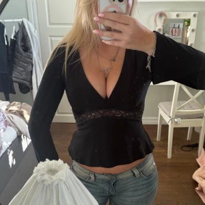 Svart omlottopp med spetsdetalj - Supersnygg svart omlottopp från Gina tricot med djup v-ringning och lång ärm. Toppen har en transparent spetsdetalj runt midjan som ger en extra touch. Materialet är mjukt och stretchigt, använd fåtal gånger🩷