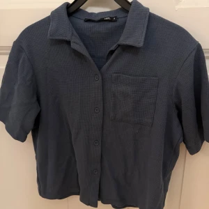 Blå kortärmad skjorta - Blå skjorta i storlek S med korta ärmar och klassisk krage. Skjortan har en strukturerad, ribbad yta och en bröstficka. Stängs med knappar framtill och har en loose passform, endast använd 1-2 gånger!